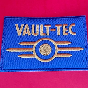 May include: A blue and gold embroidered patch with the text "Vault-Tec" and a stylized logo of a circle with three lines radiating out from it.