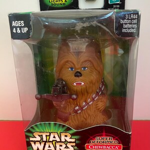 May include: A brown, furry Star Wars Chewbacca toy with blue eyes and a blaster. The toy is in a clear plastic box with a black and green Star Wars Power of the Jedi logo. The box also says "Super Deformed Chewbacca from Japan".