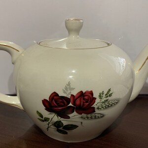 Könnte beinhalten: Weiße Teekanne mit goldener Verzierung und einem Blumendesign mit roten Rosen und Weizenähren.