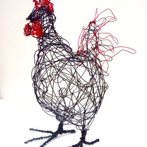 May include: A wire sculpture of a rooster. The sculpture is made of black wire with red accents on the comb and wattle. The rooster is standing on two wire legs with detailed feet. The sculpture is set against a white background.