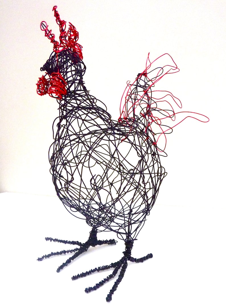 May include: A wire sculpture of a rooster. The sculpture is made of black wire with red accents on the comb and wattle. The rooster is standing on two wire legs with detailed feet. The sculpture is set against a white background.