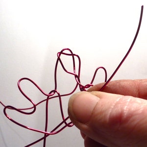 May include: Close-up of a hand manipulating a length of bright red wire. The wire is bent into looping shapes, with a single straight piece extending from the cluster. The background is plain white.