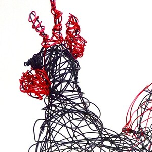 May include: A wire sculpture of a rooster, crafted from black and red wire. The rooster's comb and wattle are red, while the body is black. The sculpture is set against a white background, highlighting the intricate wirework.