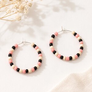 May include: A pair of hoop earrings featuring a thin silver-toned metal wire base. Each hoop is decorated with a repeating pattern of small, smooth, round beads in alternating colors of black, soft pink, and cream, creating a circular design.