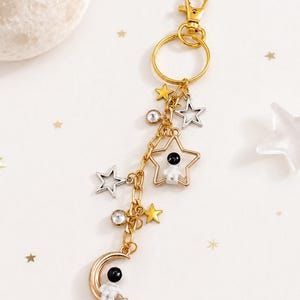 May include: A gold-toned metal keyring featuring a lobster clasp and ring, adorned with dangling charms including a crescent moon with a small astronaut, a star frame containing an astronaut, silver and gold stars, and clear round crystals.