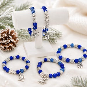 May include: Four beaded bracelets featuring alternating blue and silver glittery spheres. Two bracelets are displayed on a white T-bar stand, while two rest below. The charms include silver-toned snowflakes and snowmen figures on the bracelets.