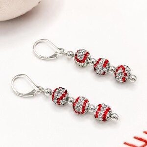 May include: A pair of drop earrings featuring silver-tone leverback hooks and three spherical beads each, designed to resemble baseballs with white and red crystal accents. The earrings are displayed alongside a partial view of a baseball.