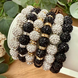 Może przedstawiać: Stos bransoletek z koralików w kolorze czarnym, srebrnym i złotym. Bransoletki wykonane są z małych, okrągłych koralików, z złotymi przekładkami. Koraliki mają błyszczący, teksturowany wygląd. Bransoletki są prezentowane na białej powierzchni.