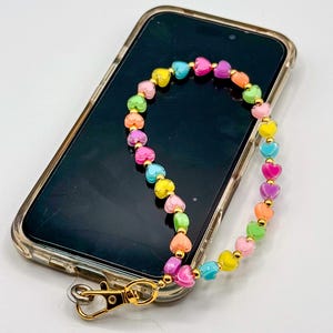 Puede incluir: Un colgante de teléfono con cuentas de colores en forma de corazón en varios colores, incluyendo rosa, amarillo, verde y azul. El colgante está sujeto a un cierre dorado y está colocado sobre un smartphone en una funda transparente.
