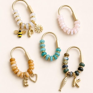 May include: Five gold-toned oval carabiner keychains adorned with colorful beads. One features a bee, letter P, and flower charm. Another has a letter E and heart charm. A third includes a fish and letter K charm. Two others have plain beads.