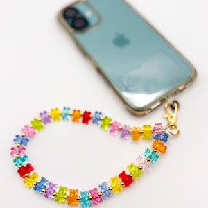 Puede incluir: Un colorido charm para teléfono con una cadena de pequeñas cuentas de ositos de goma translúcidos en varios colores, incluyendo rojo, amarillo, azul y rosa. El charm está unido a un cierre dorado y está junto a un teléfono turquesa en una funda transparente.