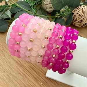 Puede incluir: Una colección de pulseras de cuentas en tonos rosas, que van desde el rosa claro hasta el fucsia intenso. Cada pulsera presenta cuentas redondas y pulidas y pequeñas cuentas espaciadoras doradas. Las pulseras se exhiben sobre una superficie blanca.