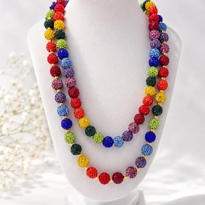 May include: A multi-strand necklace featuring round beads with a textured, glittery finish in a vibrant rainbow color palette including red, orange, yellow, green, blue, and purple, separated by small gold-toned spacers on a display bust.