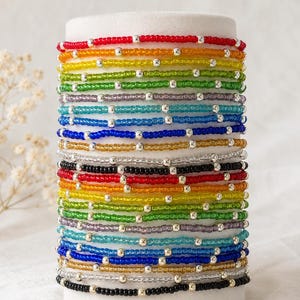 May include: A stack of multiple thin bracelets made from small, colorful glass seed beads and interspersed with tiny, reflective silver-toned metallic spacer beads. The bracelets feature a repeating rainbow color sequence including red, orange, yellow, green, purple, light blue, dark blue, gold, clear, and black.