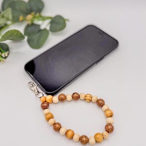 以下が含まれることがあります： 茶色とベージュの色合いの木製ビーズのリストストラップ。黒いスマートフォンに取り付けられています。ビーズはサイズと色が異なり、自然でアースカラーの美しさを生み出しています。ストラップは、銀色の留め金で電話に接続されています。