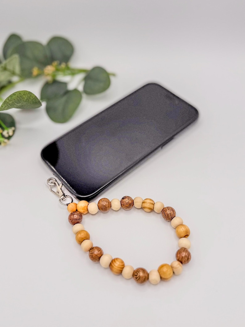 以下が含まれることがあります： 茶色とベージュの色合いの木製ビーズのリストストラップ。黒いスマートフォンに取り付けられています。ビーズはサイズと色が異なり、自然でアースカラーの美しさを生み出しています。ストラップは、銀色の留め金で電話に接続されています。