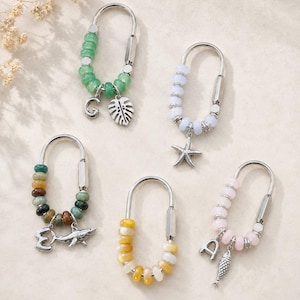 May include: Five U-shaped metal keychains featuring colorful beads and silver-toned charms. One has green beads with a letter G and leaf charm, one white beads with a starfish, one multi-colored beads with a letter E and shark, one yellow beads, and one pink beads with a letter A and fish charm.