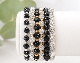May include: A set of six stacked beaded bracelets featuring alternating rows of faceted black beads, clear crystal-encrusted white beads, and smooth gold-toned metallic spacer beads, arranged in a vertical sequence on a white cylindrical display.