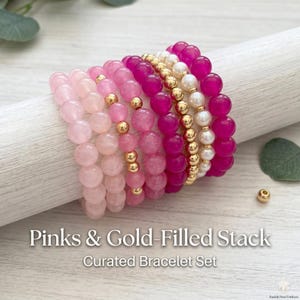 May include: A curated bracelet set featuring pink and gold-filled beads. The stack includes various shades of pink, from light to deep magenta, interspersed with gold accents and white pearl-like beads. The text "Pinks & Gold-Filled Stack" and "Curated Bracelet Set" is visible.