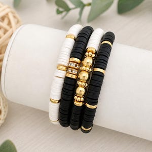 May include: A set of four stretch bracelets featuring flat, circular clay beads in alternating black and white colors. The bracelets are accented with gold-toned metal spacers, smooth gold spheres, and a central band of small, clear crystals.