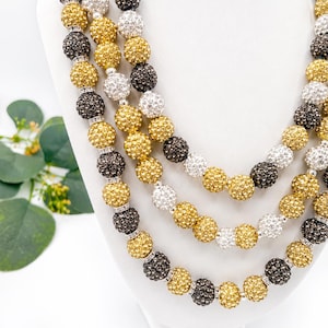 May include: A multi-strand necklace featuring round beads with a textured, sparkling finish in shades of gold, silver, and dark gray. The beads are separated by small, clear crystal-encrusted spacers, creating a layered look on a white display.