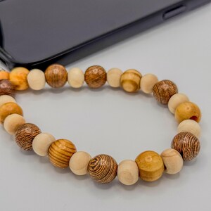 以下が含まれることがあります： 明るい茶色、ベージュ、濃い茶色の木製ビーズを組み合わせた携帯電話アクセサリー。ビーズはループ状に繋がっています。背景には黒いスマートフォンがあります。
