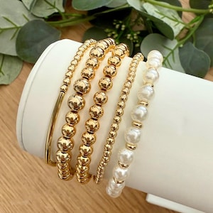 Könnte beinhalten: Eine Sammlung goldener Perlenarmbänder und ein goldener Armreif werden auf einer weißen Oberfläche präsentiert. Die Armbänder variieren im Design, darunter kleine Perlen, größere runde Perlen und Kunstperlen. Grünes Laub ist im Hintergrund zu sehen.