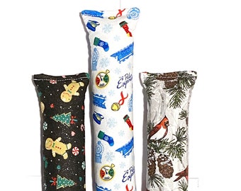 May include: Three long, rectangular cat toys with festive holiday patterns. One is black with gingerbread men and Christmas trees, another is white with blue trains and snowflakes, and the third is white with cardinals and pinecones.