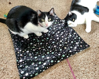 May include: Two black and white cats are on a patterned cat mat. One cat is sitting and looking at the camera, while the other is lying down. The mat is black with white and iridescent dots.