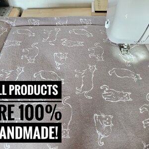 May include: A gray cat-themed fabric being sewn with a white sewing machine. The fabric has a repeating pattern of white cat illustrations. Text on the image reads: ALL PRODUCTS ARE 100% HANDMADE!