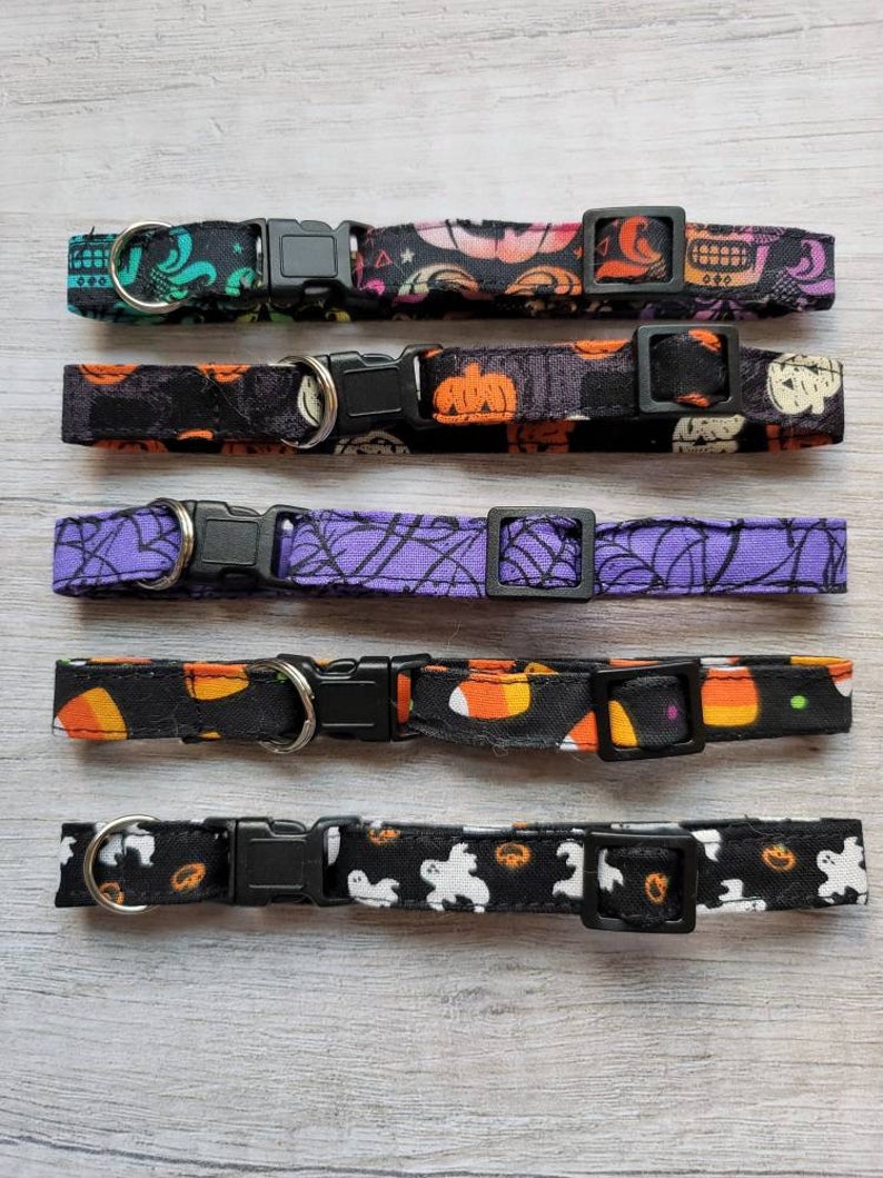 May include: Five Halloween-themed pet collars with black plastic buckles and metal D-rings. The collars feature various patterns, including skulls, pumpkins, spiderwebs, candy corn, and ghosts. Each collar is approximately 1 inch wide.