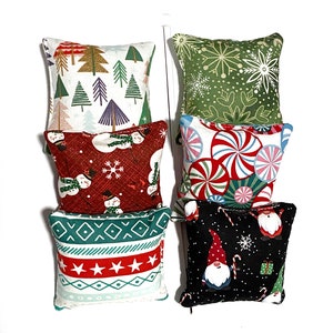Può includere: Una collezione di piccoli cuscini quadrati a tema natalizio. I cuscini presentano vari motivi festivi, tra cui alberi di Natale, fiocchi di neve, pupazzi di neve, bastoncini di zucchero e gnomi. La tavolozza dei colori comprende rosso, verde, bianco e nero.