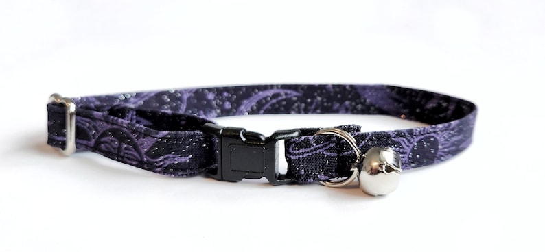 May include: A black and purple patterned cat collar with a silver bell and a black plastic buckle. The collar is made of fabric and has a repeating design. The bell is attached with a silver ring.