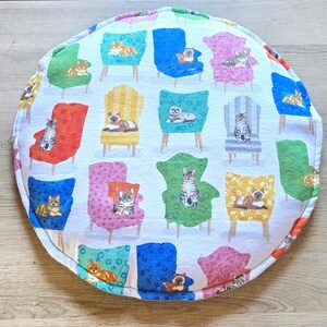 May include: Round cat bed with a white background and a repeating pattern of colorful armchairs, each with a cat. The chairs are in shades of red, blue, green, yellow, and pink. The cats are in various poses.