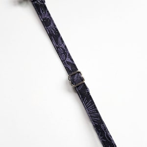 May include: A black cat collar with a repeating purple sun and moon pattern. The collar has a silver-colored bell, a metal buckle, and a black plastic clasp. The collar is designed for a cat.