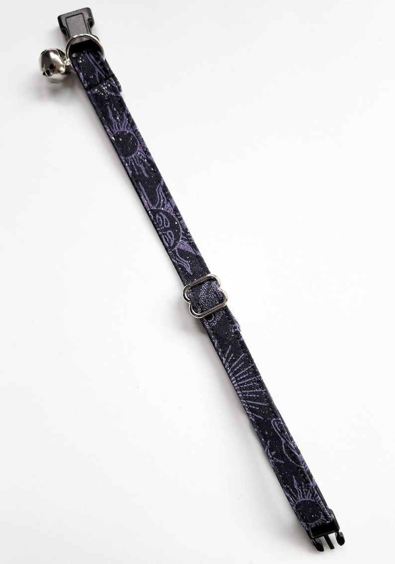May include: A black cat collar with a repeating purple sun and moon pattern. The collar has a silver-colored bell, a metal buckle, and a black plastic clasp. The collar is designed for a cat.