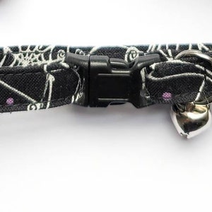 May include: Black cat collar with a Halloween-themed pattern of white spiderwebs, orange stars, and purple dots. The collar has a black plastic buckle and a silver bell.