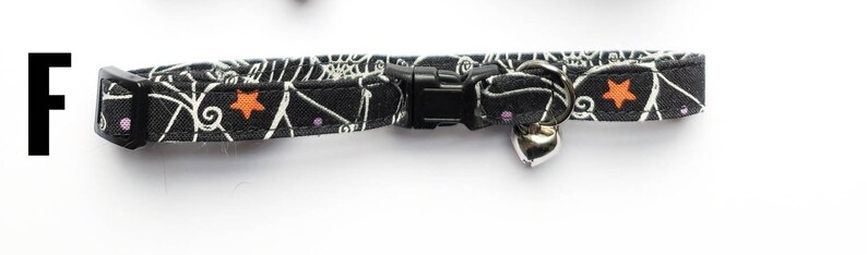 May include: Black cat collar with a Halloween-themed pattern of white spiderwebs, orange stars, and purple dots. The collar has a black plastic buckle and a silver bell.