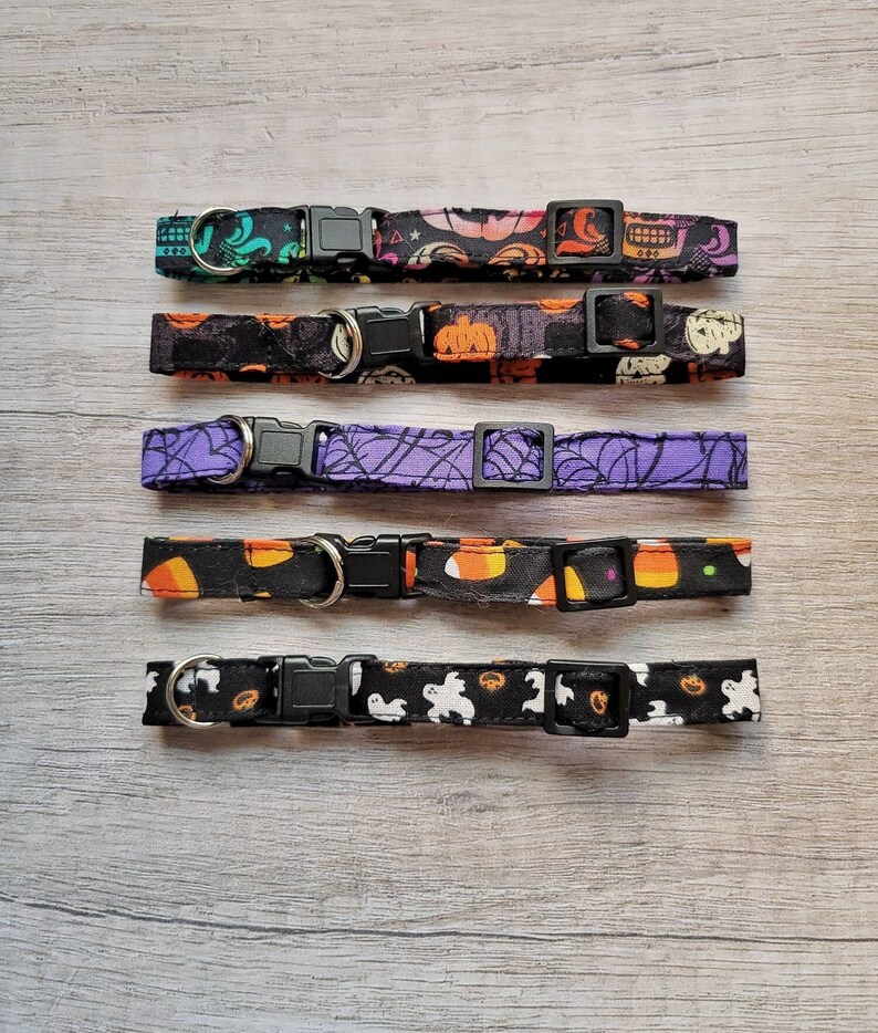 May include: Five Halloween-themed pet collars with black plastic buckles and D-rings. The collars feature various patterns, including ghosts, pumpkins, spiderwebs, and candy corn, in colors like black, orange, purple, and teal. The collars are laid out on a light wood surface.