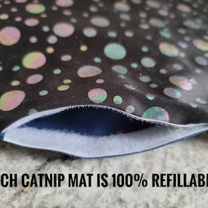 May include: Close-up of a black catnip mat with iridescent, colorful polka dots. The mat has a white opening and the text "EACH CATNIP MAT IS 100% REFILLABLE!"
