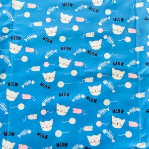 May include: A blue pet bed with a repeating pattern of white cat faces, fish bones, pink mice, and the word "MEOW" in black. The bed has a quilted design.