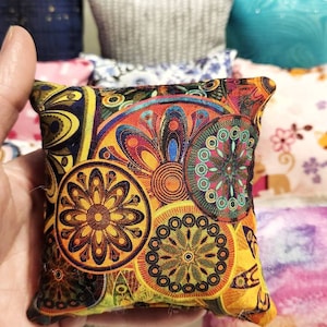 K&ouml;nnte beinhalten: Ein kleines, quadratisches Kissen mit einem lebendigen, farbenfrohen Blumenmuster in Gelb-, Orange-, Blau- und Gr&uuml;nt&ouml;nen. Das Kissen wird in einer Hand gehalten, im Hintergrund sind weitere Kissen zu sehen.