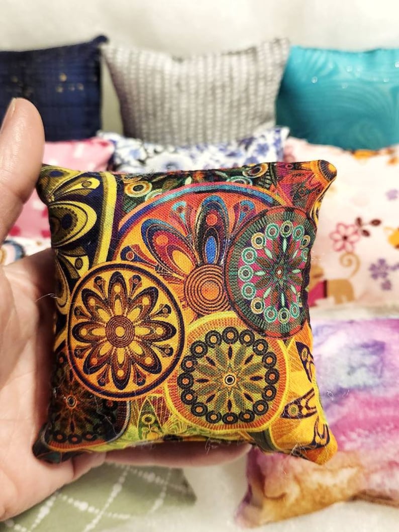 K&ouml;nnte beinhalten: Ein kleines, quadratisches Kissen mit einem lebendigen, farbenfrohen Blumenmuster in Gelb-, Orange-, Blau- und Gr&uuml;nt&ouml;nen. Das Kissen wird in einer Hand gehalten, im Hintergrund sind weitere Kissen zu sehen.