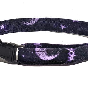 May include: A dark purple cat collar with a celestial pattern of moons and stars. The collar has a black plastic buckle, a silver-colored bell, and a metal loop for size adjustment.