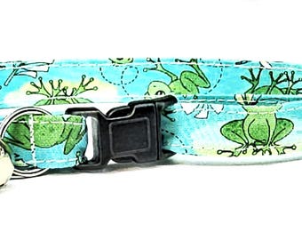 Op de afbeelding: Een turquoise kattenhalsband met een herhalend patroon van groene kikkers en witte bloemaccenten. De halsband heeft een zwarte plastic gesp, een zilveren metalen D-ring en een kleine zilveren bel.