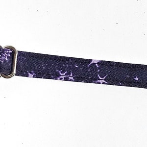 May include: A black strap with a repeating pattern of white moons and stars. The strap has a silver metal adjuster. The strap is likely for a bag or other accessory.