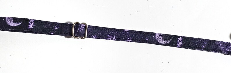 May include: A black strap with a repeating pattern of white moons and stars. The strap has a silver metal adjuster. The strap is likely for a bag or other accessory.