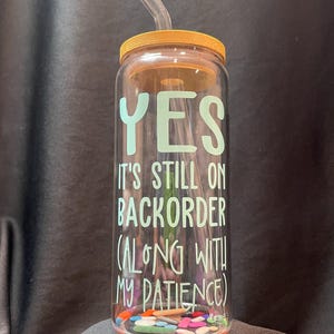 Könnte beinhalten: Eine klare Glaswasserflasche mit Bambusdeckel und Strohhalm. Die Flasche hat ein grünes und weißes Design mit dem Text "YES IT'S STILL ON BACKORDER (ALONG WITH MY PATIENCE)".