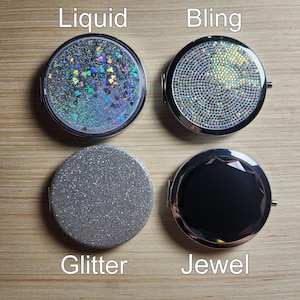 May include: Four compact mirrors with different designs. The top left mirror has a liquid-like design with iridescent glitter. The top right mirror has a bling design with clear rhinestones. The bottom left mirror has a silver glitter design. The bottom right mirror has a black jewel design.