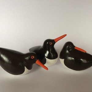 May include: Three ceramic oystercatcher figurines, each with a black and white body and an orange beak. The birds are positioned in a group, with one facing forward and the other two facing sideways.